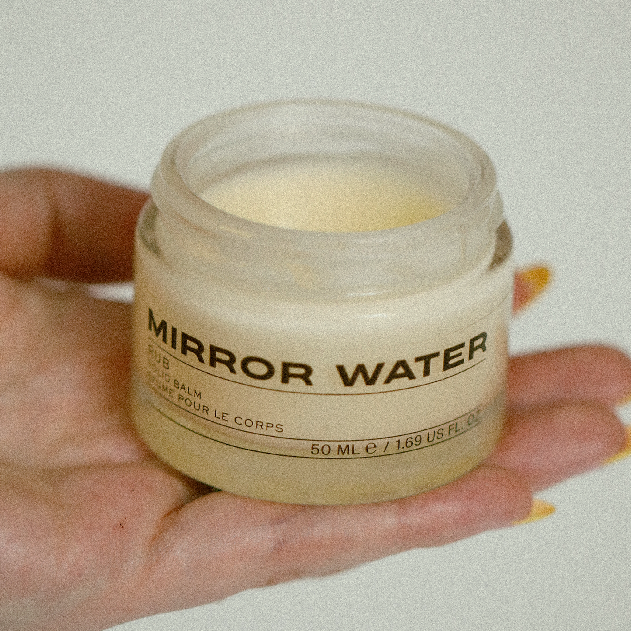50ml jar of MIRROR WATER in models hand. The jar is open showing cream coloured product.