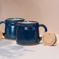 Two fairtrade dark blue ceramic mugs with fairtrade tags on a white surface with botanical shadows.