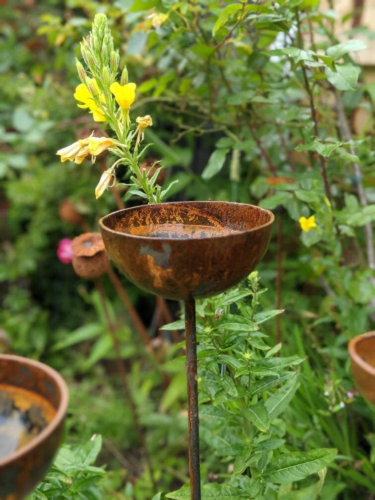 Close up focussing on one Rain Catcher amongst a group of three. Background is green foliage and flowers. The attractive rust metal patina is clearly shown.