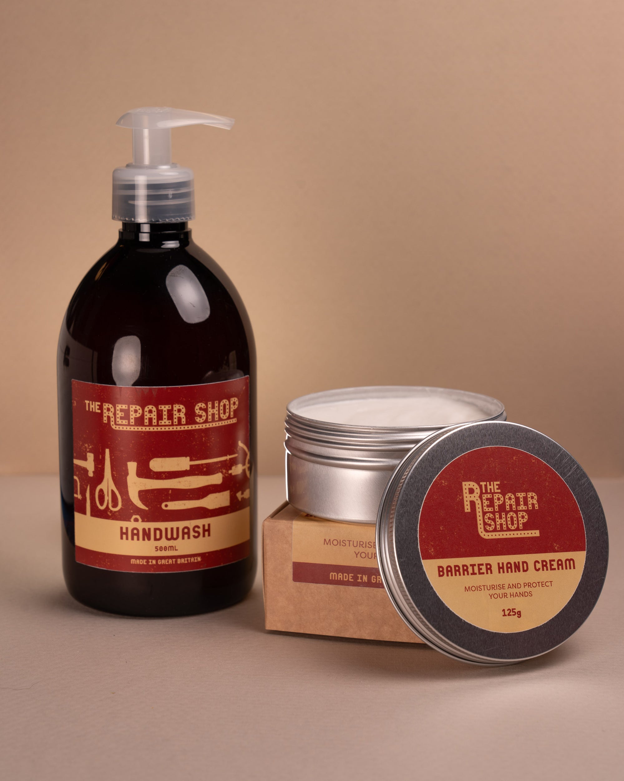 A bottle of The Repair Shop handwash beside a silver pot of The Repair Shop Barrier hand cream a styled in a studio shot on beige backdrop. The  pot of hand cream is open showing white hand cream. 