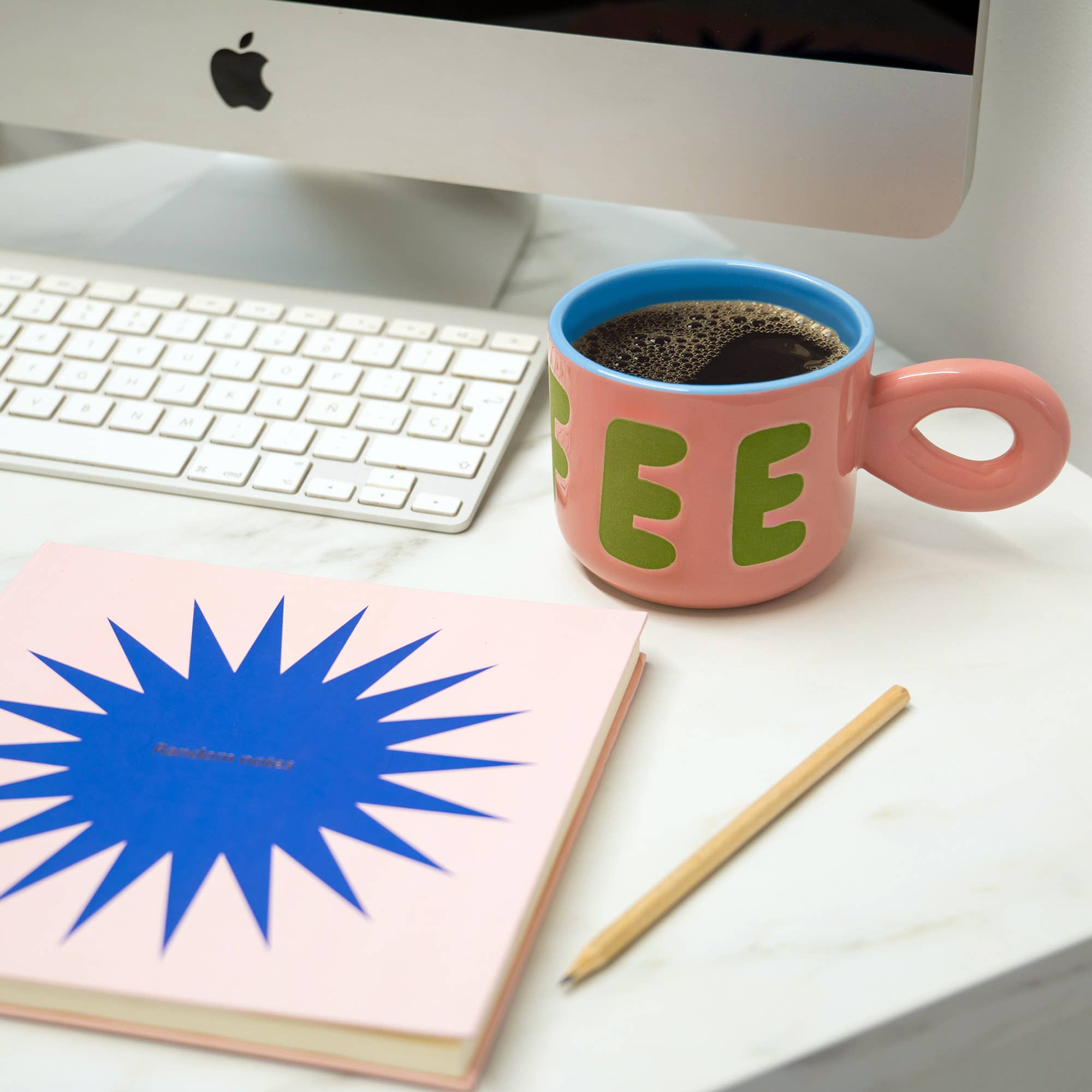 Pink mug with 'COFFEE' text, notebook, and pencil on a desk with a computer.