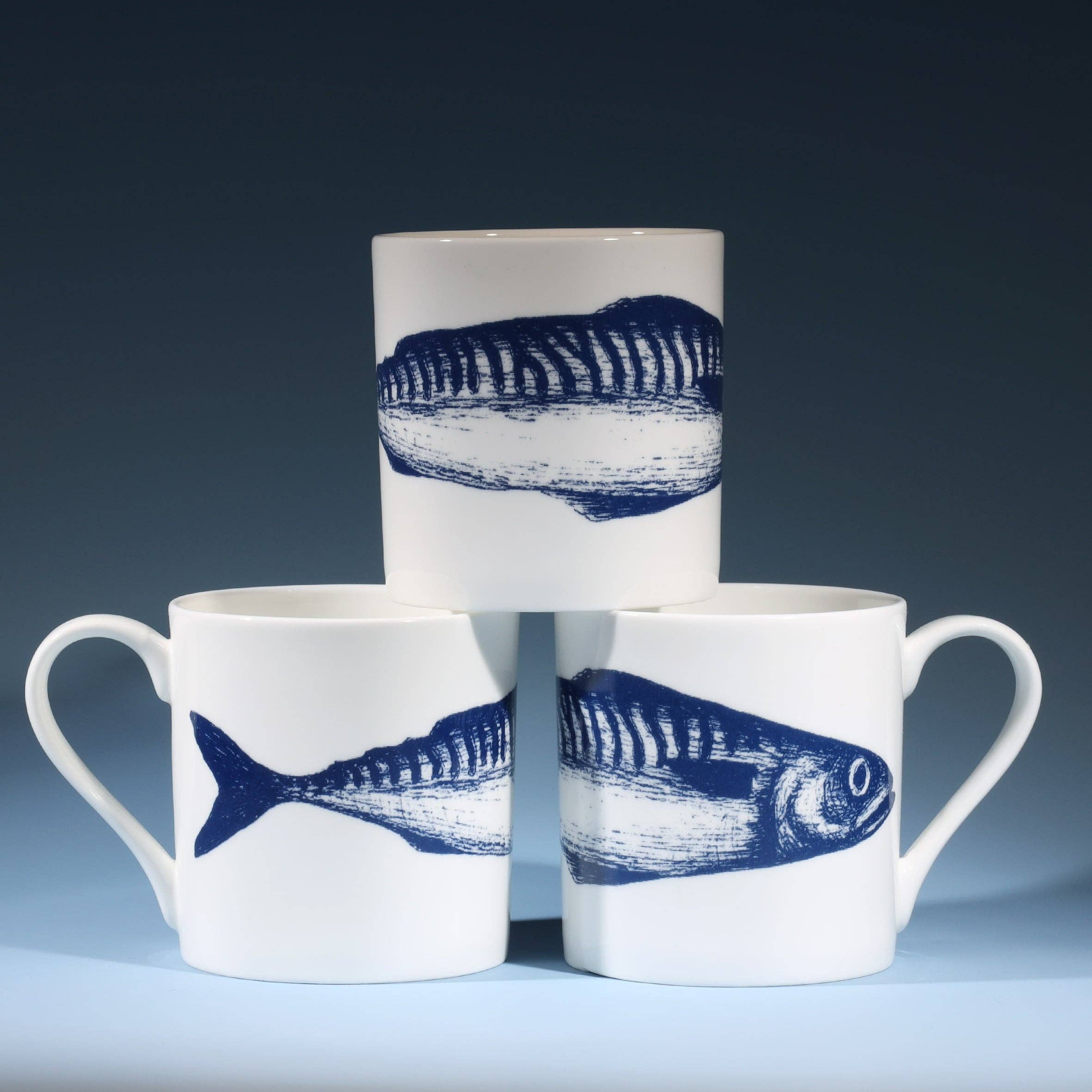 Stack of three Bone China Mackerel Mugs showing all sides of fish illustration. Studio shot on inky blue background. Great gift!
