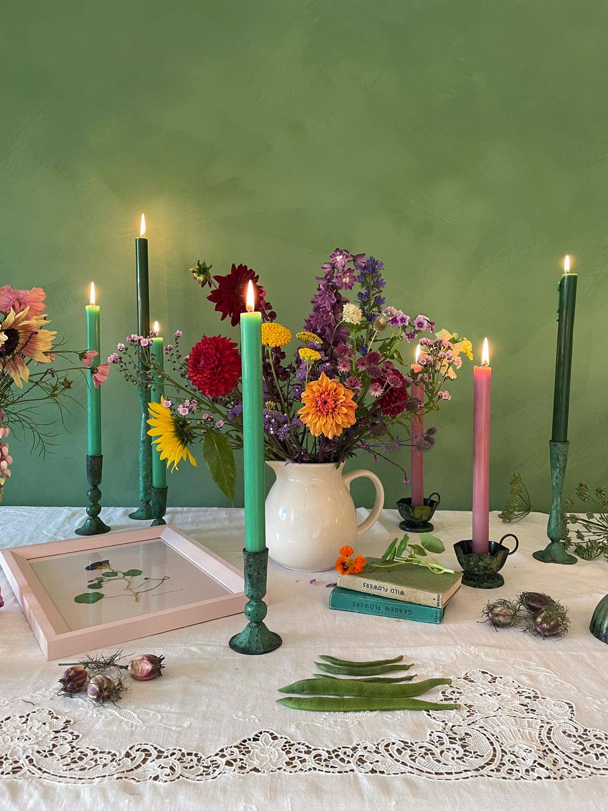 Collection of verdigris candlesticks of varying sizes on a table top with vase of summer flowers .Colourful flowers and candles against summery green backdrop. Handmade verdiris candlesticks - perfect for table top styling.