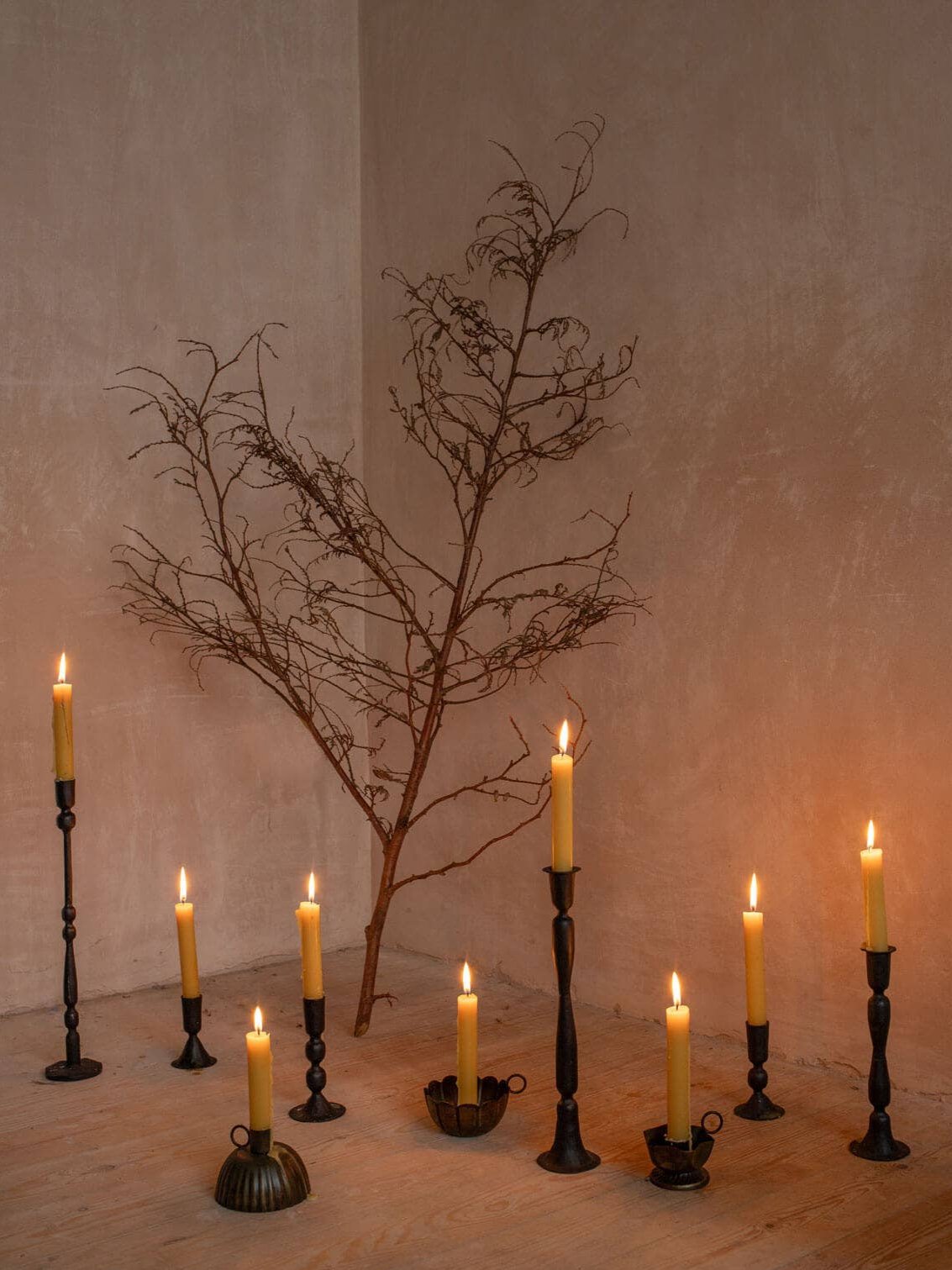 9 candlesticks of varying heights and sizes all with lit white candles, arrange on a floor with a branch in the background propped against plaster coloured wall.